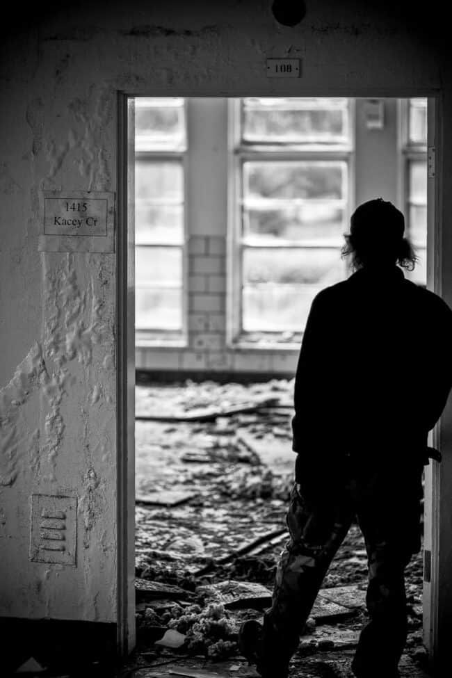 A black-and-white photo of a person in silhouette standing in the doorway of an abandoned, debris-strewn room, seen from a hallway with numbered room placards