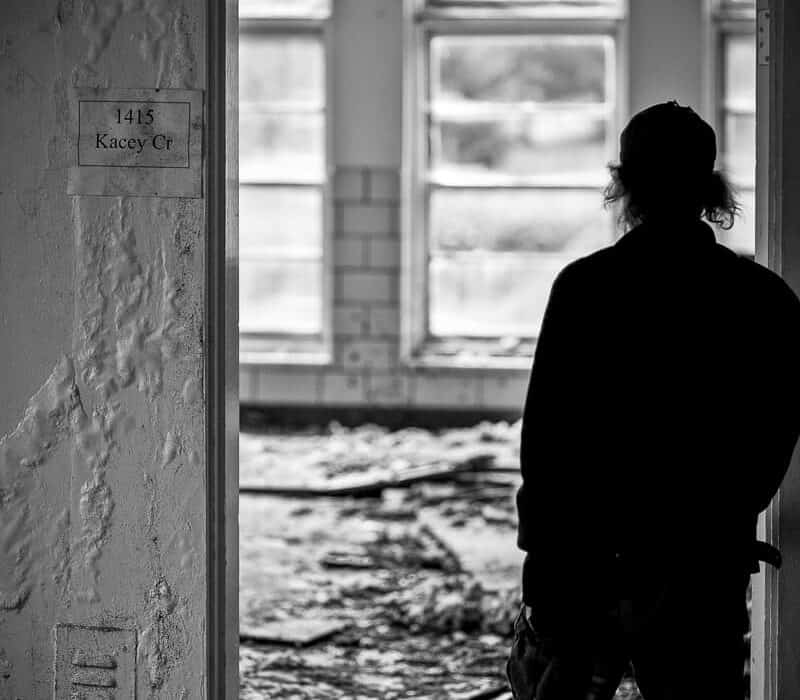A black-and-white photo of a person in silhouette standing in the doorway of an abandoned, debris-strewn room, seen from a hallway with numbered room placards