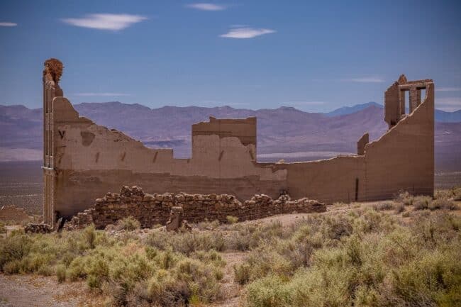 The crumbling adobe-brick ruins of a ghost town building, with two chimney-like towers still standing against desert mountains