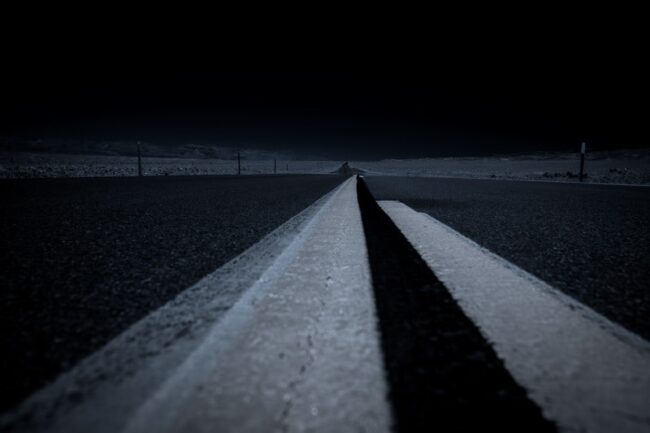 A moody black-and-white long-exposure photo of an empty highway at night, with double yellow lines glowing under headlights, in Death Valley National Park, California