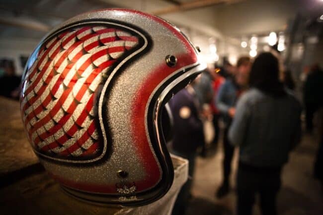 A close-up of a vintage motorcycle helmet with a red and silver flame-scale paint design, a blurred crowd in the background