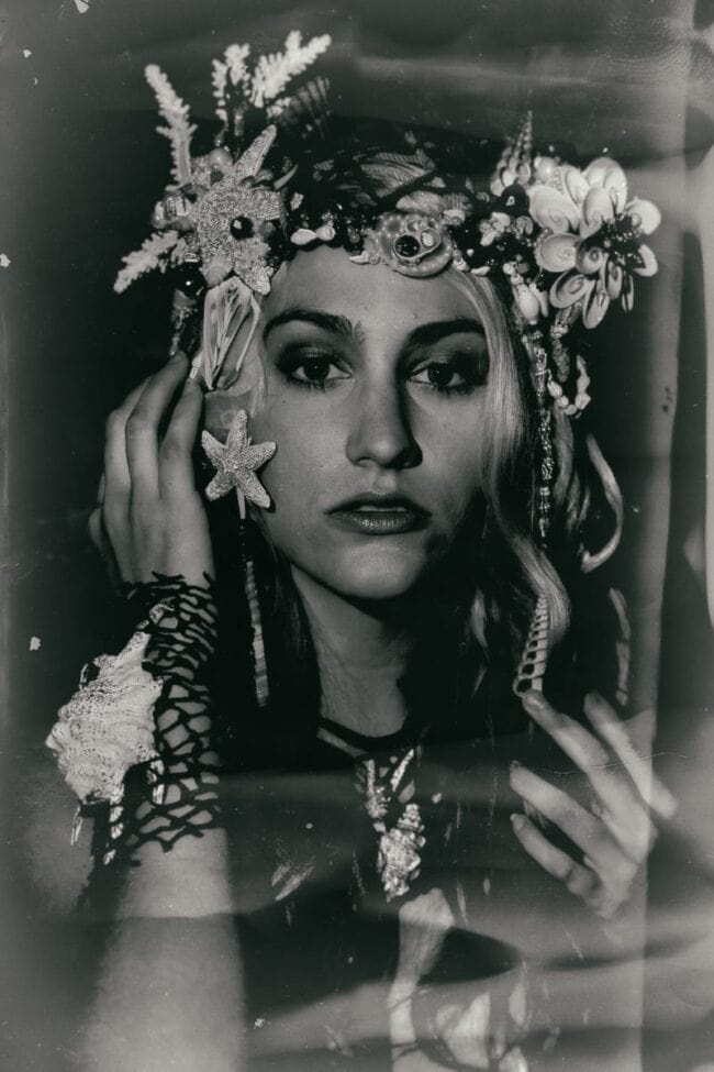 A vintage-style wet-plate black-and-white portrait of a woman wearing an elaborate headpiece and jewelry made of starfish, shells, and coral, hand raised to her face