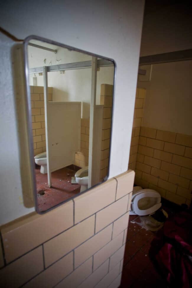 A cracked mirror reflecting a row of toilet stalls in an abandoned, decaying bathroom, tile walls and debris on the floor