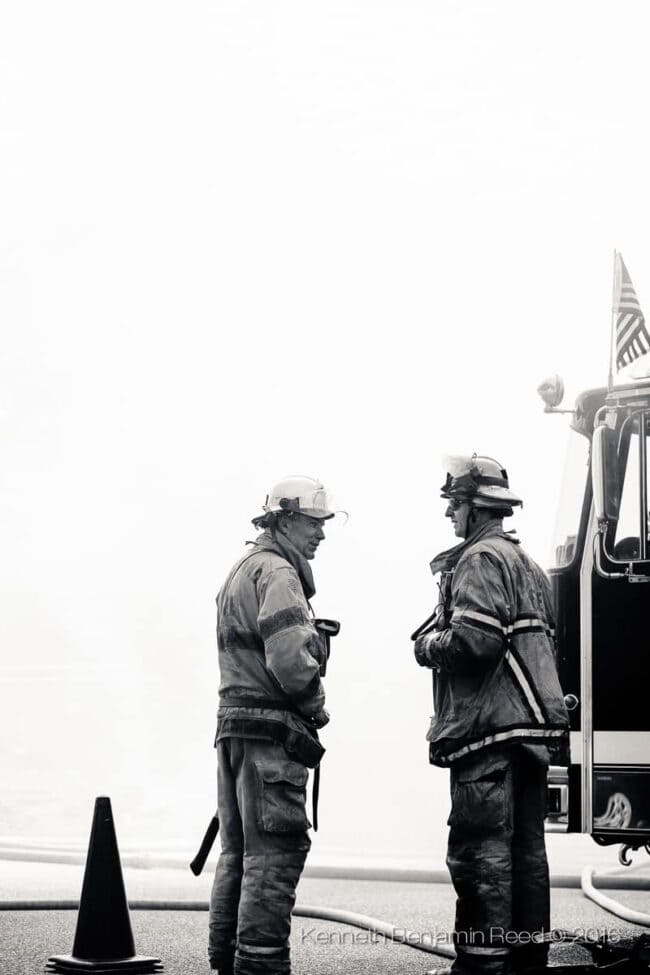 A black-and-white photo of two firefighters in full gear talking beside a fire truck flying an American flag, on a hazy day