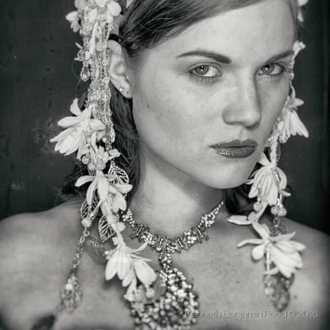 A black-and-white close-up portrait of a woman wearing an ornate floral headpiece and layered necklace, with a direct serious gaze