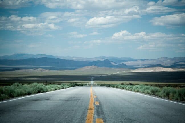 An empty two-lane highway with a bright yellow center line stretching toward distant desert mountains under a partly cloudy sky