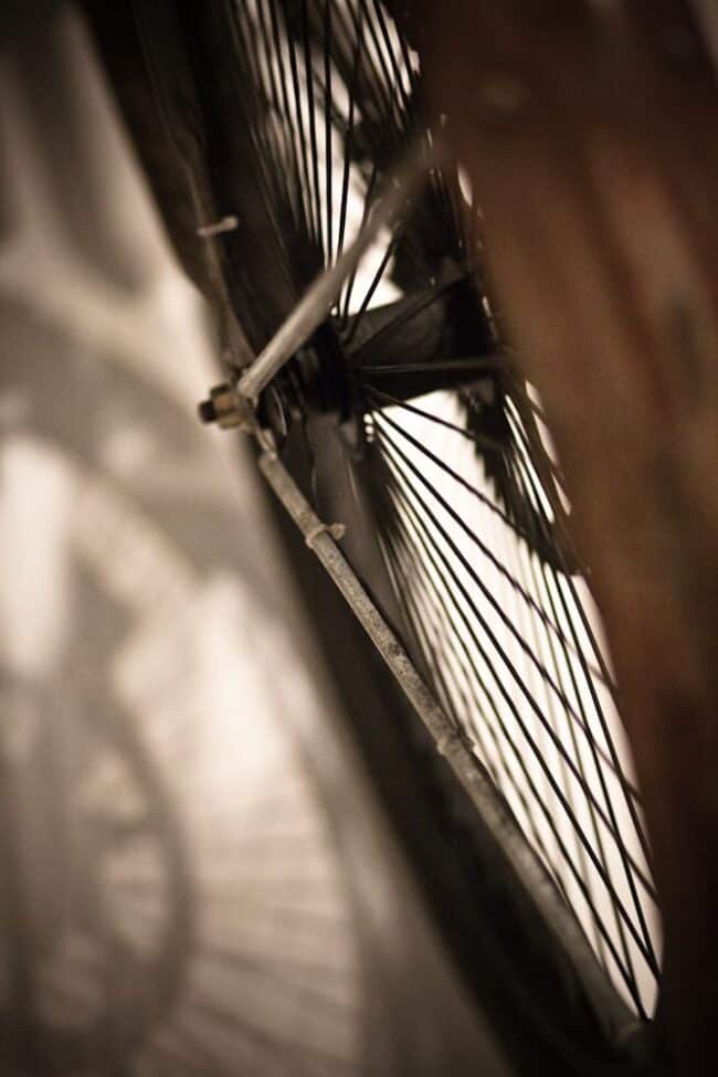 A close-up sepia-toned photo of a vintage bicycle wheel with wire spokes, backlit against a wood floor with the shadow of an ornate clock face