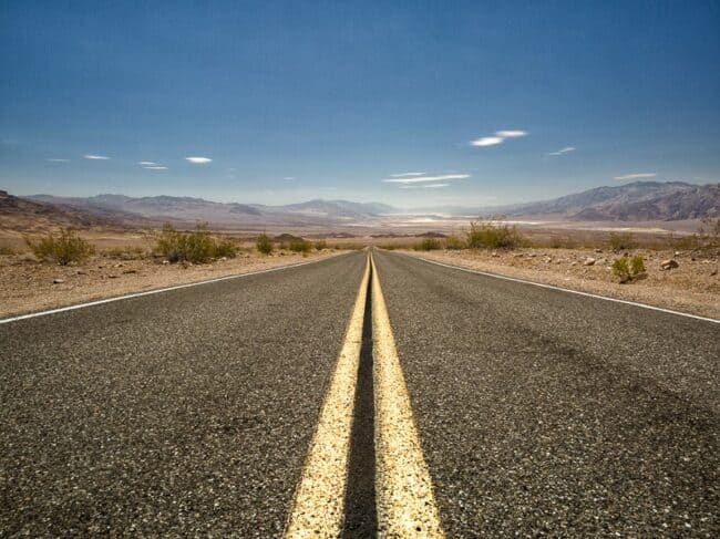 An empty two-lane highway with a bright double yellow center line stretching toward distant desert mountains near the Death Valley, California and Nevada border