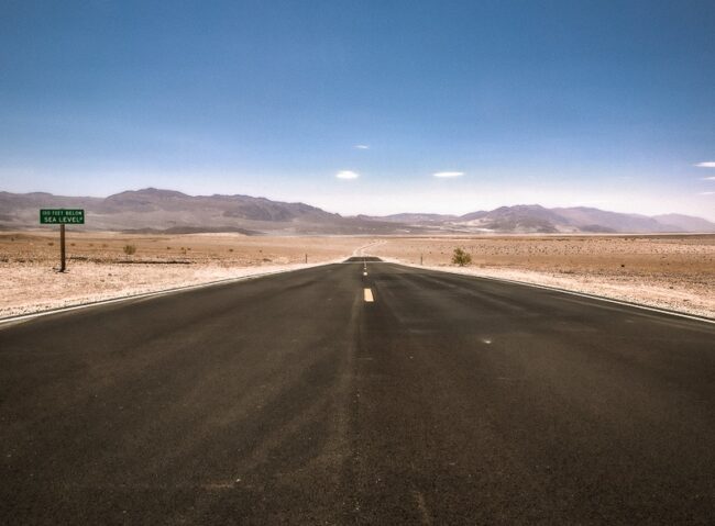 An empty desert highway with a small green road sign noting the elevation below sea level in Death Valley National Park, California, mountains toward Nevada in the distance