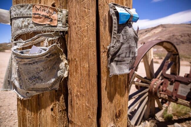 Weathered denim shorts pockets nailed to a wooden post as a makeshift trail register, with a rusted wagon wheel and desert hills behind