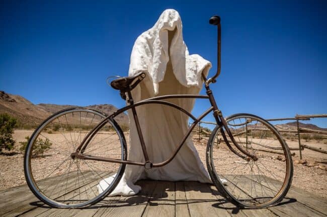 A vintage bicycle draped in a white sheet styled to look like a ghost, parked on a wooden boardwalk in the desert