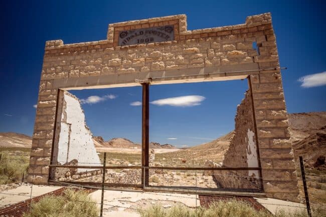 The stone facade of a ruined general store building, with a faded sign reading "R. Porter 1906" above an empty window frame overlooking the desert