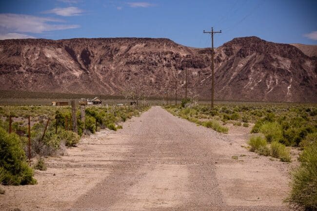 A dirt road leading toward distant mountains, passing old buildings and a utility pole, in a Nevada ghost town