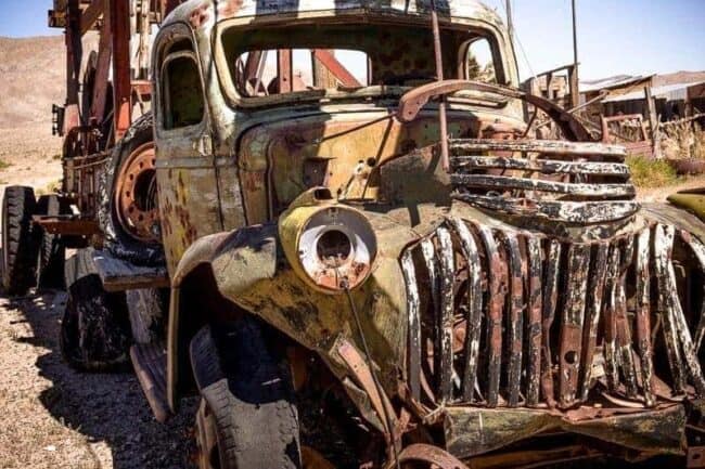 The rusted, weathered front end of an abandoned vintage truck with a skeletal grille and peeling green paint, in a desert junkyard