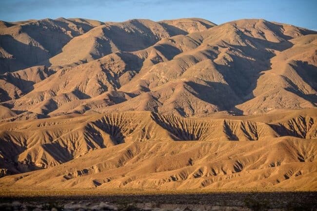 Sun-lit desert mountains with deeply eroded ridges and canyons, seen from a distance