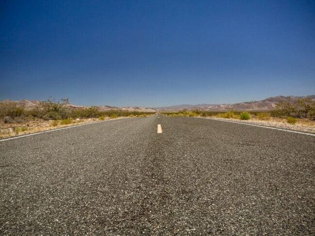 An empty desert highway stretching to the horizon under a clear blue sky, low scrub brush lining the road
