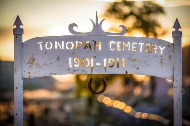 A weathered white cemetery sign reading "Tonopah Cemetery 1901-1911" with a horseshoe hanging from it, backlit by sunset