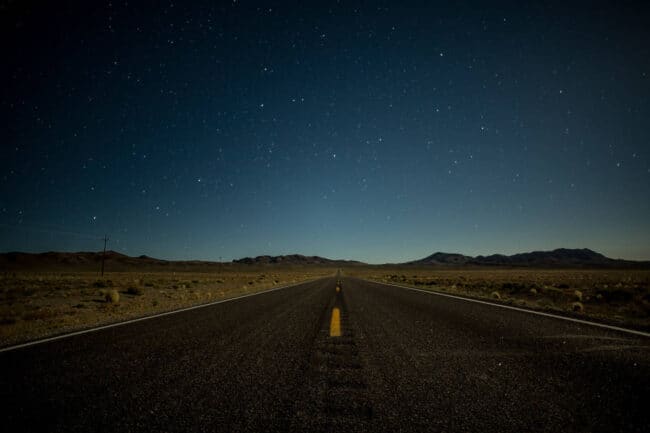 An empty desert highway at night under a star-filled sky, the yellow center line glowing faintly in the darkness