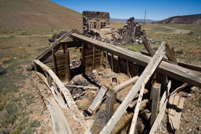 An aerial view of a collapsed wooden roof structure next to ruined stone walls in a desert ghost town, with a dirt road curving into the distance