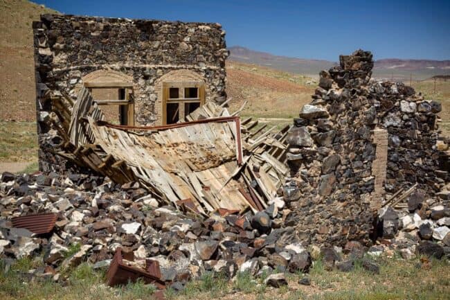 A partially collapsed stone ruin with two arched window openings still visible, walls crumbling into rubble under a wide desert sky