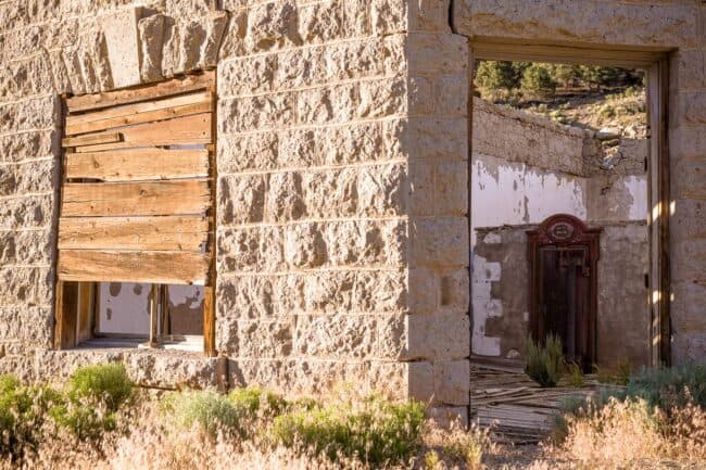 An abandoned stone building in a ghost town, with a boarded-up window and an open doorway revealing a rusted antique safe inside