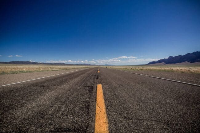 An empty two-lane highway stretching to the horizon under a bright blue sky, with desert mountains in the distance