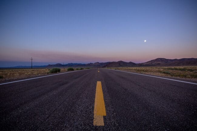 An empty two-lane highway stretching toward the horizon at dusk, with the moon visible above distant mountains