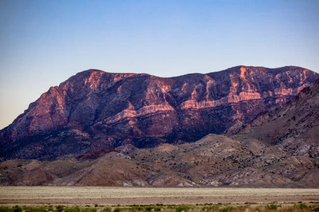 A reddish desert mountain ridge lit by warm evening light, with a flat sagebrush plain in the foreground