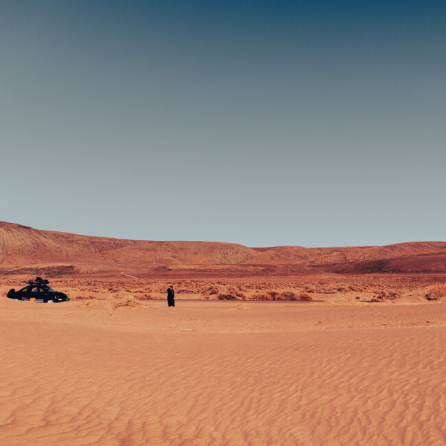 A desert landscape with a vintage car and a lone figure in dark clothing standing on rippled sand dunes, distant mountains under a clear sky