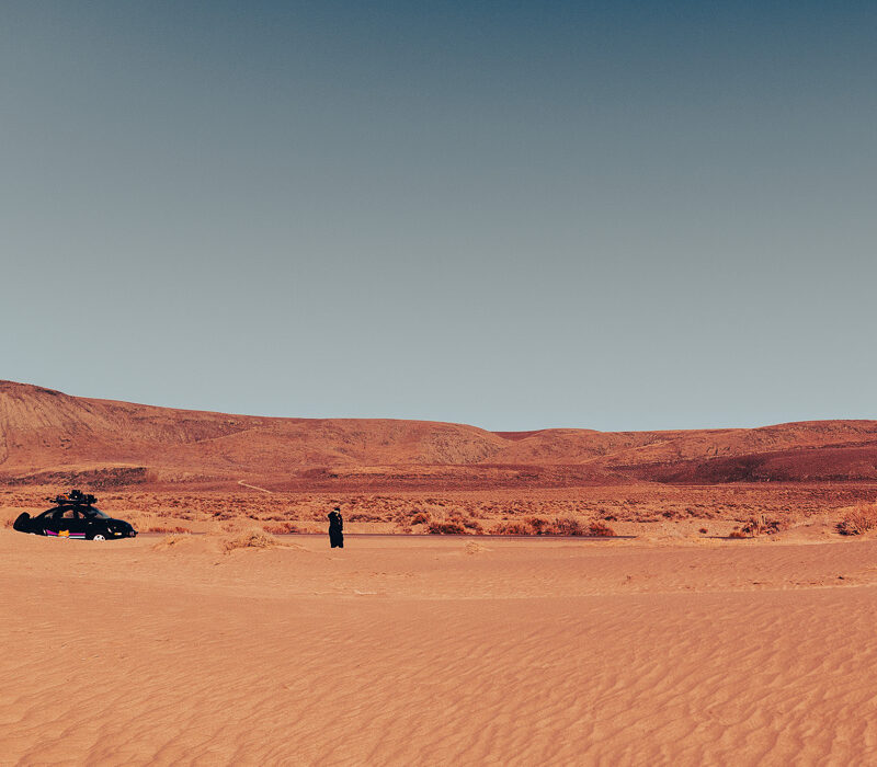 A desert landscape with a vintage car and a lone figure in dark clothing standing on rippled sand dunes, distant mountains under a clear sky