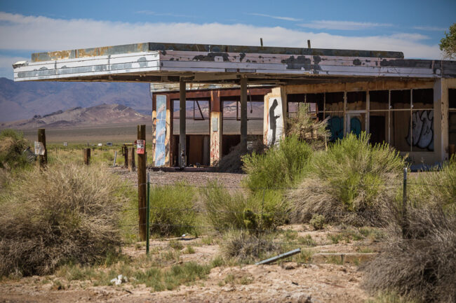An abandoned gas station with a graffiti-covered facade and collapsed canopy roof, desert mountains in the background