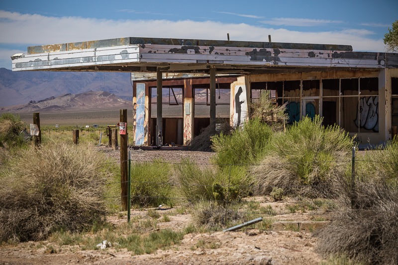 An abandoned gas station with a graffiti-covered facade and collapsed canopy roof, desert mountains in the background