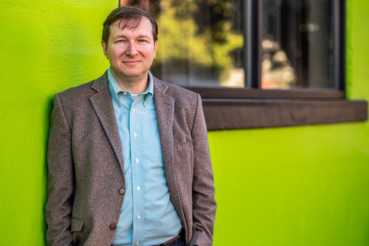 Man in a gray tweed blazer and light blue shirt leaning against a bright green wall, color