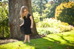 Young girl in a black dress walking on sunlit grass near a large tree